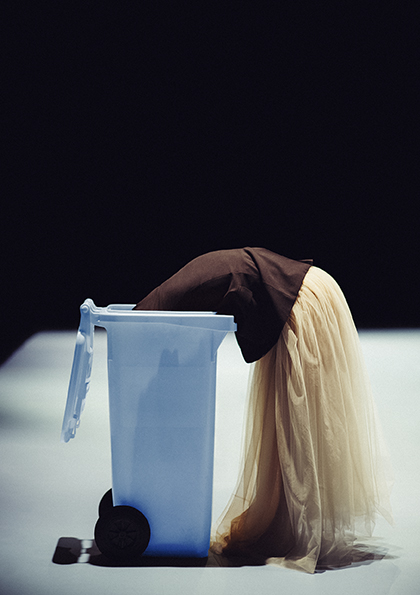Een persoon staat voorovergebogen in een lichtblauwe vuilnisbak. Ze draagt een lichtgekleurde, vloeiende rok. De foto heeft dramatische belichting met een donkere achtergrond en een verlichte vloer, wat een theatrale of artistieke sfeer creëert.