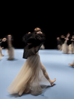 Dansers in lange, vloeiende rokken bewegen over een lichtblauw toneel, hun bewegingen vervaagd door de snelheid, tegen een zwarte achtergrond.