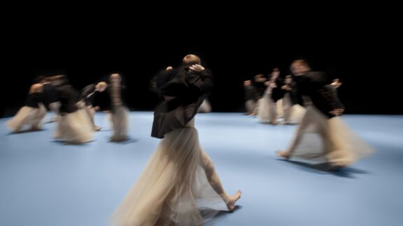 Dansers in lange, vloeiende rokken bewegen over een lichtblauw toneel, hun bewegingen vervaagd door de snelheid, tegen een zwarte achtergrond.