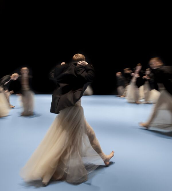 Dansers in lange, vloeiende rokken bewegen over een lichtblauw toneel, hun bewegingen vervaagd door de snelheid, tegen een zwarte achtergrond.