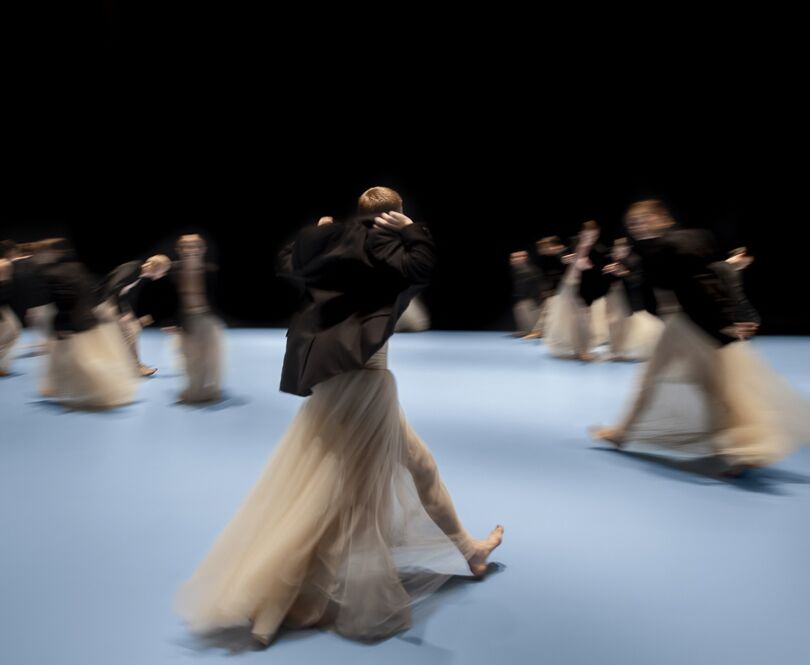 Dansers in lange, vloeiende rokken bewegen over een lichtblauw toneel, hun bewegingen vervaagd door de snelheid, tegen een zwarte achtergrond.