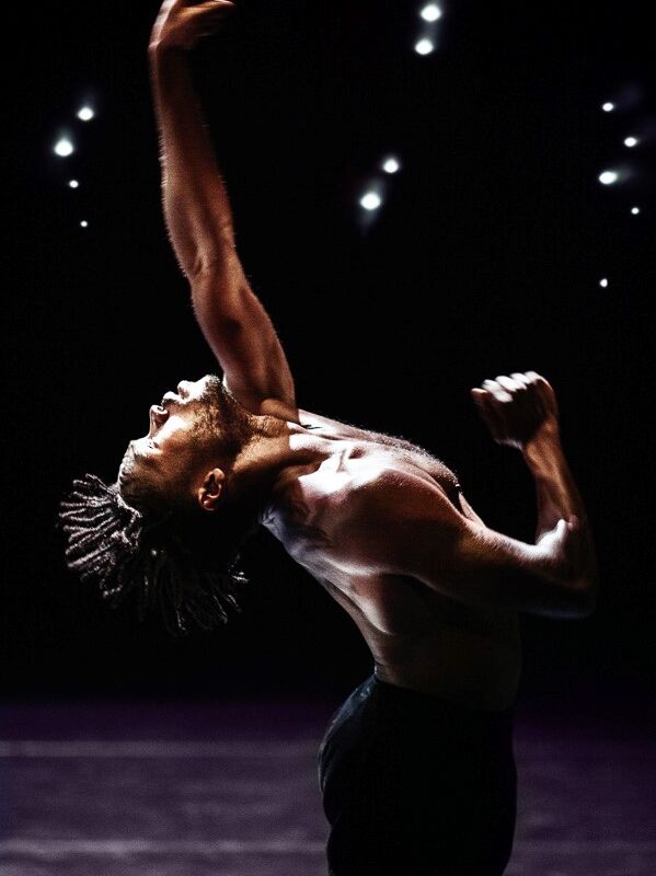 A contemporary dancer performs on stage under dramatic lighting, with arms raised and body arched backward in an expressive pose. The dancer has dreadlocks and is shirtless, captured mid-movement against a dark background with stage lights visible above.