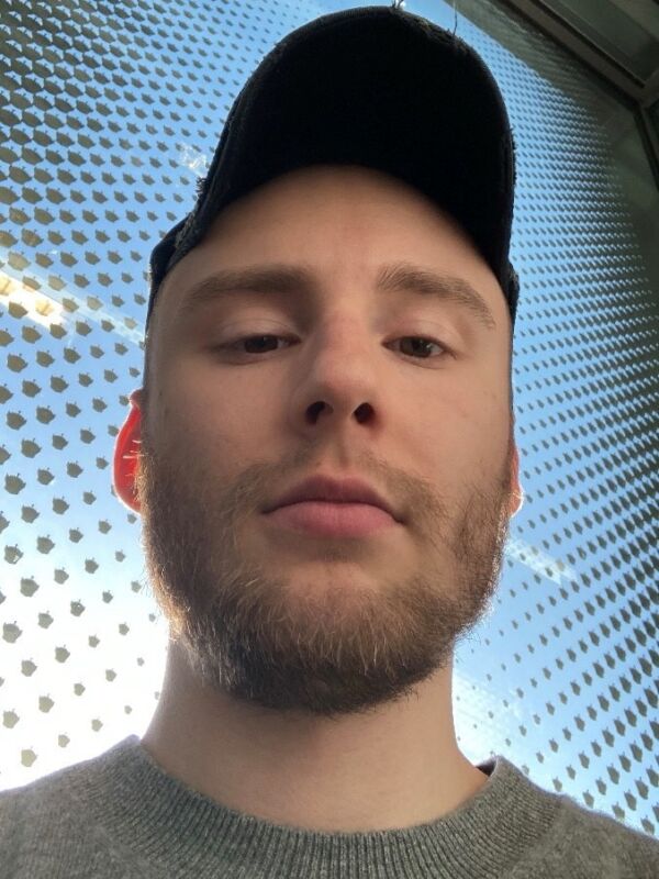 Selfie van een jongeman met een baard, een zwarte pet en een grijze trui. De foto is van onder genomen, met een patroon van stippen op het raam en blauwe lucht op de achtergrond.