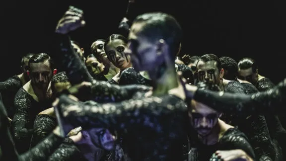 A group of dancers with intense expressions and dark face paint move closely together in a powerful formation