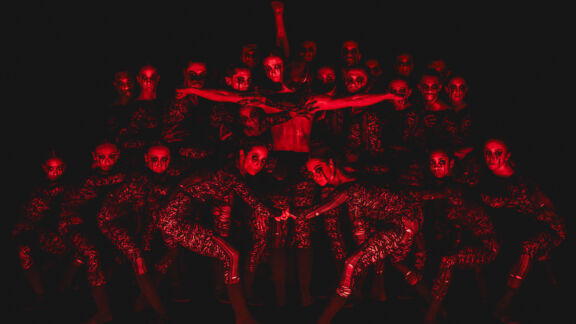 Dancers in patterned costumes pose under red light with intense expressions and dramatic arm movements.