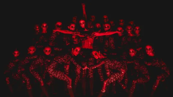 Dancers in patterned costumes pose under red light with intense expressions and dramatic arm movements.