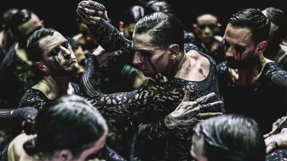 Dancers in intricate black costumes perform intensely under dim lighting, their expressions focused and movements dynamic, conveying a dramatic tension.