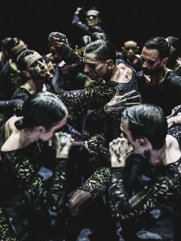 Dancers in intricate black costumes perform intensely under dim lighting, their expressions focused and movements dynamic, conveying a dramatic tension.