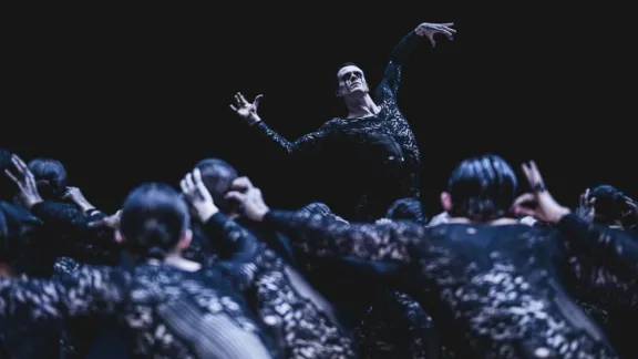 Dancers in intricate, dark costumes perform on stage. One central dancer strikes a dramatic pose, arms raised, conveying intensity and elegance.