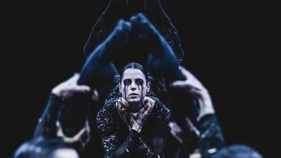 A dancer is lifted above a group, framed by raised arms and legs, with intense eye makeup and a solemn expression