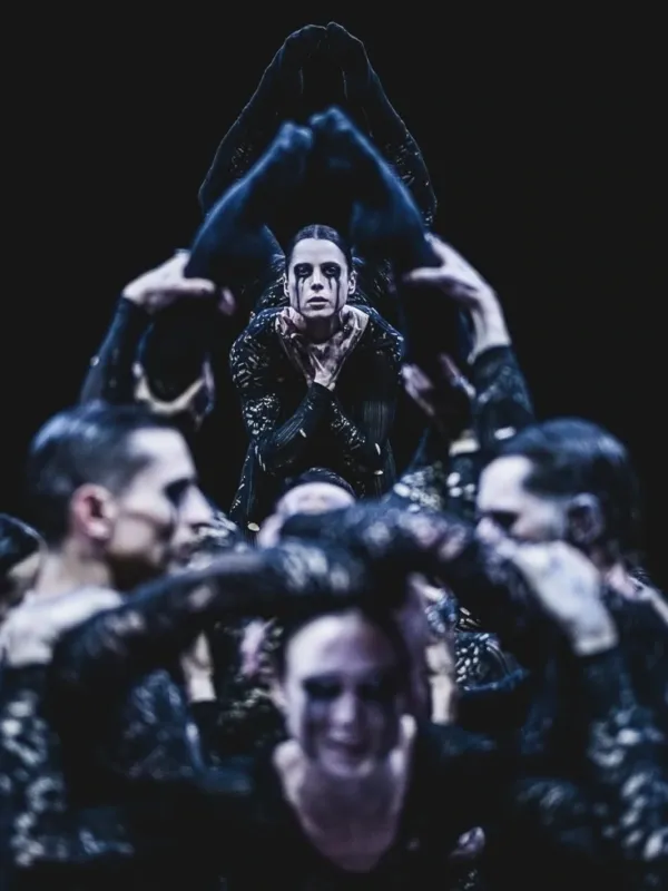 A dancer is lifted above a group, framed by raised arms and legs, with intense eye makeup and a solemn expression