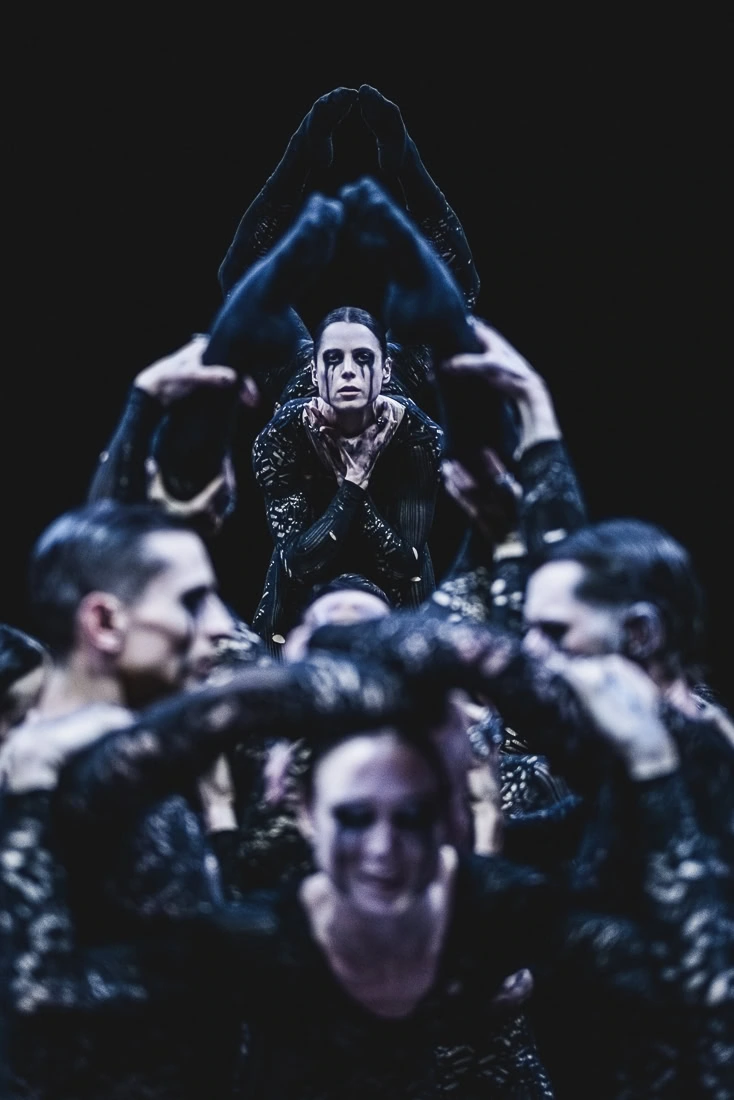 A dancer is lifted above a group, framed by raised arms and legs, with intense eye makeup and a solemn expression