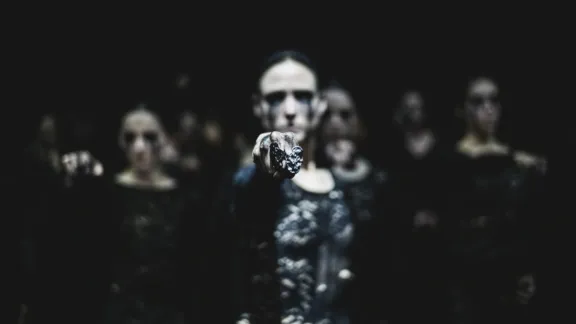 Dancers in dark lace costumes stand in a dimly lit space. One extends a hand forward, creating a mysterious and intense atmosphere.