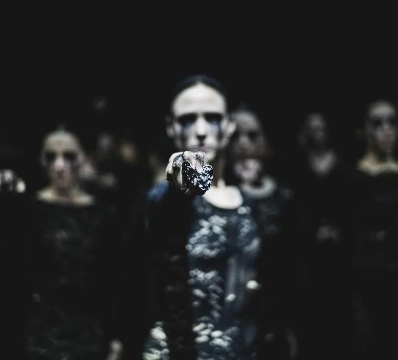 Dancers in dark lace costumes stand in a dimly lit space. One extends a hand forward, creating a mysterious and intense atmosphere.