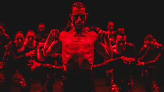 A shirtless dancer in red lighting stands confidently in a group, highlighting muscular form and intensity. The image conveys a dramatic, powerful atmosphere.