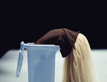 Een persoon staat voorovergebogen in een lichtblauwe vuilnisbak. Ze draagt een lichtgekleurde, vloeiende rok. De foto heeft dramatische belichting met een donkere achtergrond en een verlichte vloer, wat een theatrale of artistieke sfeer creëert.