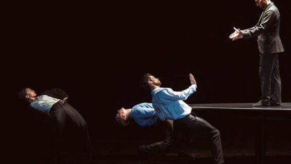 Four dancers in suits: three lean back dramatically while one stands on a table facing them with outstretched hands