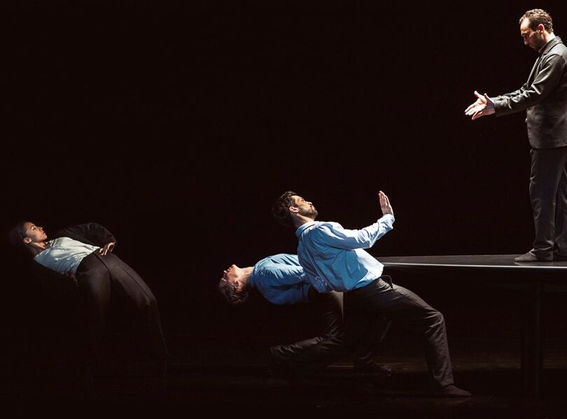 Four dancers in suits: three lean back dramatically while one stands on a table facing them with outstretched hands