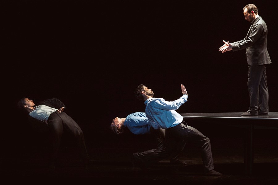 Four dancers in suits: three lean back dramatically while one stands on a table facing them with outstretched hands