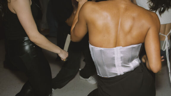 People dancing closely together at a party or club; a woman in a silver strapless top and black pants is in the center of the frame, facing away from the camera, with others dancing around her in dim lighting.