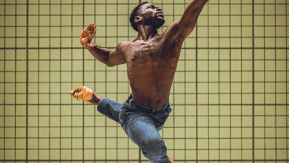 Een mannelijke danser springt krachtig door de lucht met gespreide armen en gebogen benen, tegen een achtergrond van een gele betegelde wand. Boven hem hangt een digitale klok met de tijd 20:10.