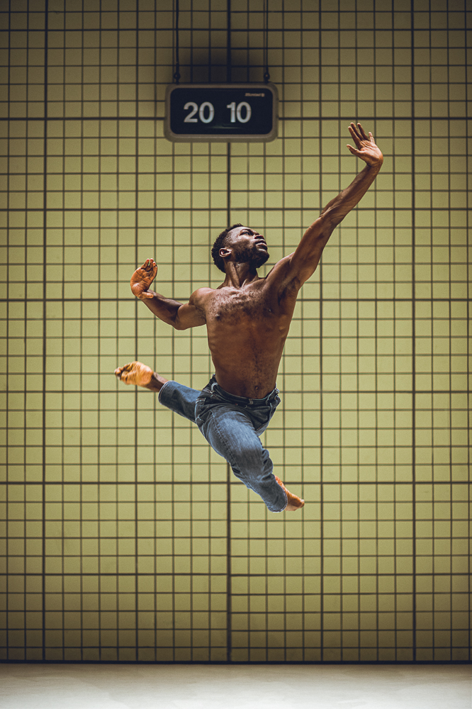 Een mannelijke danser springt krachtig door de lucht met gespreide armen en gebogen benen, tegen een achtergrond van een gele betegelde wand. Boven hem hangt een digitale klok met de tijd 20:10.